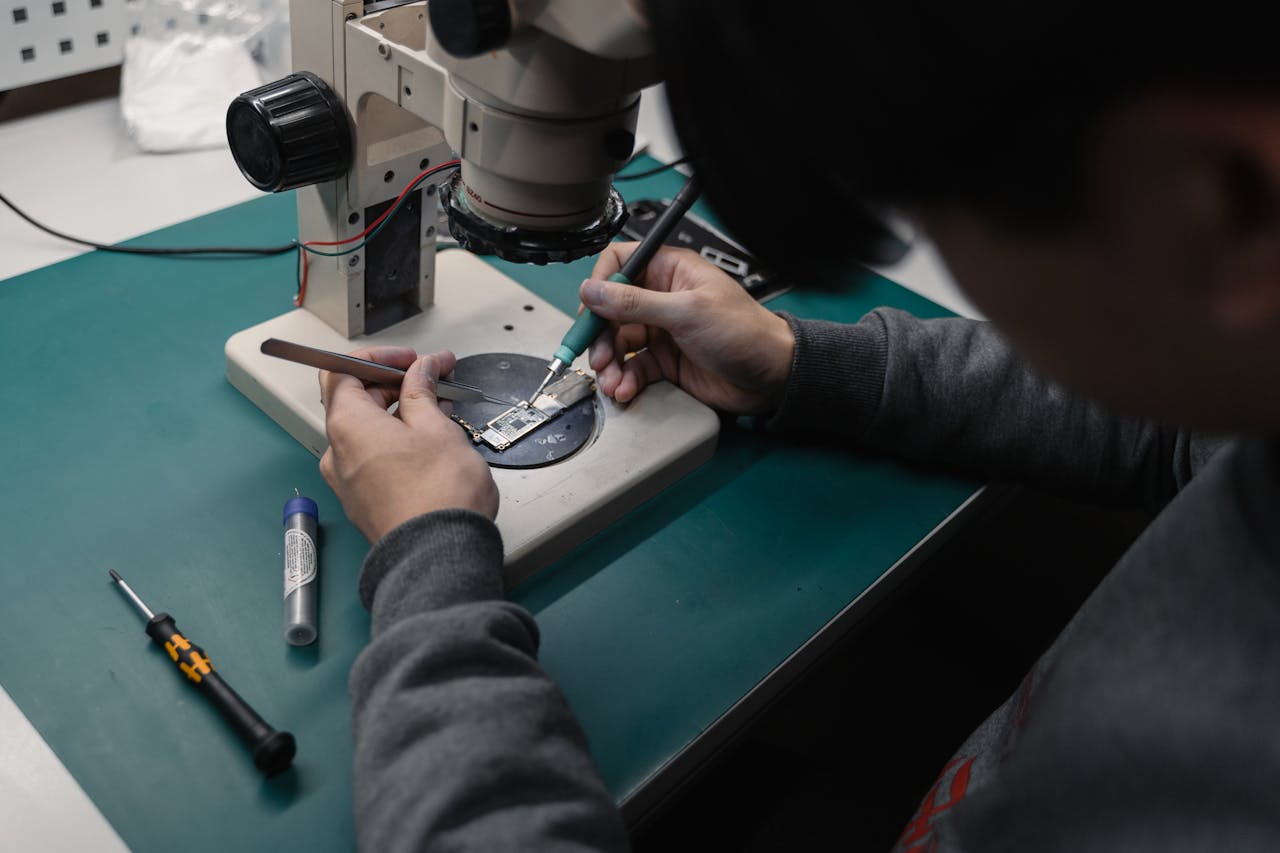 Technician using a microscope to repair a broken electronic board with precision tools.